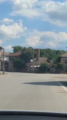 The video shows a view from a vehicle traveling down a paved road in a village on a sunny day with fluffy white clouds in a blue sky. On the left side of the road, there are several old, partially dilapidated buildings with red tile roofs and balconies. Some windows are broken or boarded up. Trees and vegetation grow around and on some of the structures. On the right side of the road, there are more buildings, some appearing to be in better condition with white walls and red roofs, while others are also weathered and run-down. There are utility poles and wires visible throughout the scene. The road curves to the left and then straightens out. Traffic signs and barriers are also present. The overall impression is of an older village with a mix of decaying and maintained structures.