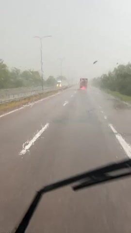 The video shows a view from inside a vehicle driving on a highway during a heavy rainstorm. The road is wet and appears to have standing water in some areas, creating spray as vehicles pass. Multiple vehicles are visible, including a white truck in the distance on the left, a red truck ahead in the right lane, and a silver car in the left lane that appears to be hydroplaning or losing control, splashing water significantly. The visibility is reduced due to the rain and overcast sky, with trees lining the sides of the highway. A windshield wiper is visible in the foreground, periodically clearing the view.