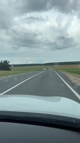 A dashcam perspective shows a two-lane asphalt road stretching into the distance under a cloudy sky. The road is bordered by green fields. A white car is visible in the distance on the right lane, moving away from the camera. The foreground shows the hood of a white vehicle.