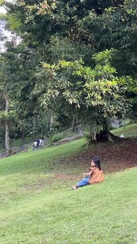 A young girl with long, dark hair sits on a grassy slope in a park-like setting. She is wearing a brown shirt and blue jeans. In her hands, she holds a green bottle. The girl appears to be relaxed and engaged with her surroundings. The background features lush green trees, with dappled sunlight filtering through the leaves, suggesting it is daytime, possibly late afternoon due to the warm lighting. Further in the background, a wooden fence and a few other people are visible, hinting at a public space. The overall atmosphere is peaceful and serene. The location appears to be a well-maintained outdoor area, with a gentle incline of the lawn. There is no visible text in the media file.