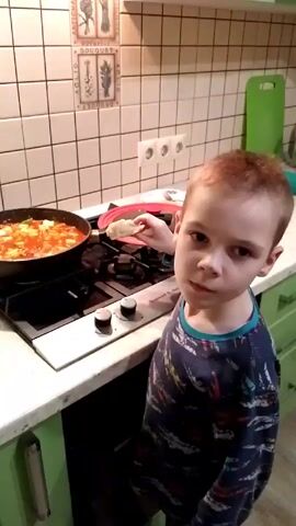 Here's a description of the video:

A young boy with short red hair stands in a kitchen, looking towards a stovetop where a frying pan sits with food cooking in it. He wears a dark blue and multicolored long-sleeved shirt. The kitchen features white-tiled walls with decorative art pieces. The stove is white with black burners and gray knobs. The countertops are light-colored, and the cabinets are green. The boy holds a piece of food in his hand and appears to be smelling the food in the pan. There is also a green cutting board behind the stove.