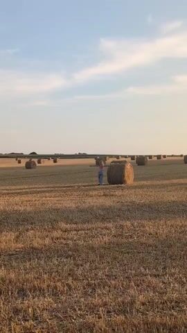 The video shows a person standing in a field of hay bales. The person is wearing a pink top and blue jeans. The field is wide and stretches into the distance, with many hay bales scattered throughout. The sky is blue with some white clouds, and the sun appears to be setting, casting a warm glow over the scene. The person is facing away from the camera and appears to be looking out at the field. The scene suggests a peaceful and rural setting.