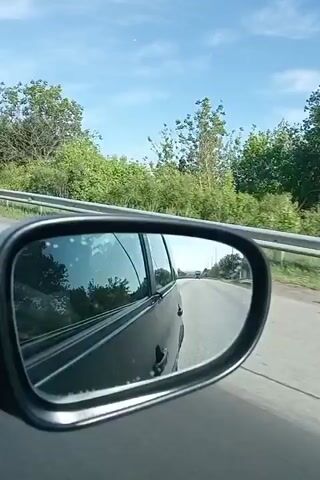 The video, shot from inside a moving car, focuses on the rearview mirror. The mirror reflects the road behind, a black car body, and the passing scenery. This includes green trees, clear blue sky with light clouds, and occasional roadside buildings. The car appears to be driving on a highway or major road with a guardrail. The video shows movement indicating the car is traveling.