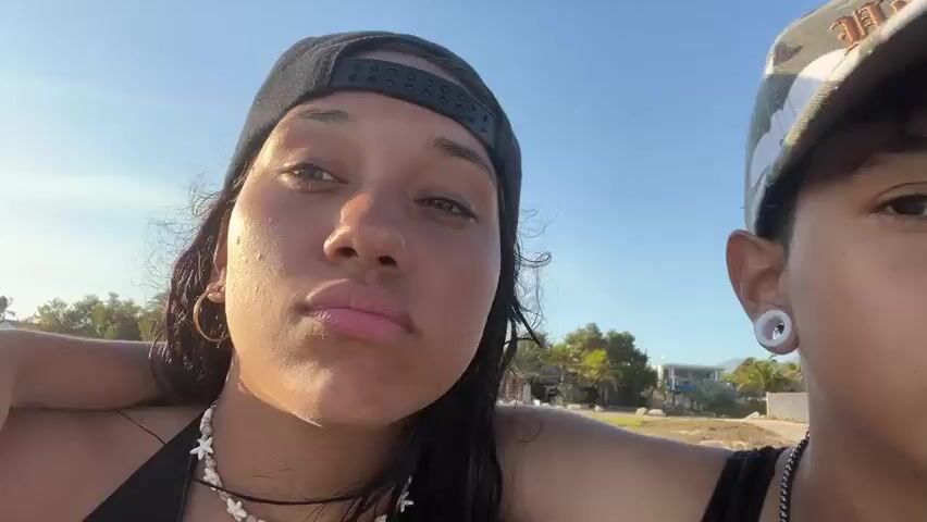 Two young women stand close together outdoors, presumably on a beach. The woman on the left wears a black baseball cap, a black bikini top, and a beaded necklace. She has dark hair, a nose ring, and gold hoop earrings. She makes a kissing face at the camera. The woman on the right wears a gray baseball cap and a black tank top. She has short hair and a silver necklace. She sticks her tongue out at the camera. The background shows sand, some trees, and buildings. The sky is clear and blue.