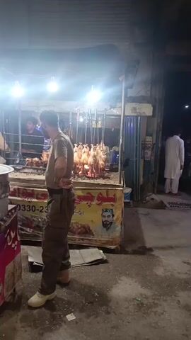 A video depicts a man standing next to a barbecue stand, where whole chickens are vertically skewered over a charcoal fire. The man is wearing a brown t-shirt, dark pants, and light-colored shoes. He has short dark hair and a beard. The barbecue stand is decorated with yellow and red signage, featuring an image of a man in a traditional cap. Behind the stand, a blue container and chairs are visible. The setting appears to be an outdoor urban area, possibly at night, with streetlights illuminating the scene. In the background, another person wearing a white garment can be seen walking away. The camera captures the scene with a somewhat shaky perspective.