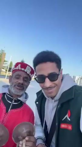 Two men are outdoors, possibly in a coastal area. The older man on the left is dressed in traditional Moroccan attire: a red and white embroidered robe and a red hat with white embellishments. He holds a pair of dark-colored, rounded musical instruments, possibly castanets, and plays them. The younger man on the right wears sunglasses, a gray hoodie, and a green jacket with a patch that says “ORIGIN.” He looks at the camera with a slight smile. The background shows a paved area, greenery, streetlights, and buildings in the distance. The sky is clear and blue. The men appear to be interacting amiably, possibly in a street performance or cultural exchange setting.