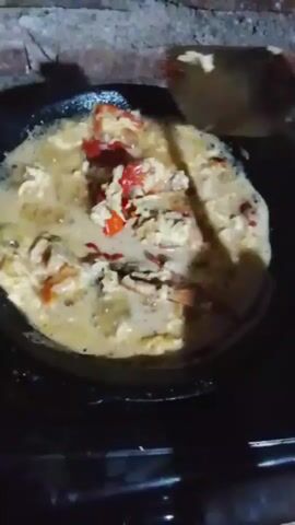A close-up shot shows a black pan filled with a mixture of crab and scrambled eggs being stirred with a wooden spatula. The crab appears to be cooked, with its red shell pieces visible among the yellow scrambled eggs and a light-colored sauce. The pan is placed on a dark surface, possibly a stove, and the background is out of focus, revealing a textured brick wall. The lighting is dim, highlighting the food in the pan. The overall scene suggests a home-cooked meal being prepared.