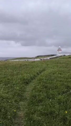 This media file captures a scenic view of the Irish coastline, likely near Dunkineely, Ireland. The foreground is dominated by a lush, green grassy field with a faint path winding through it. Small yellow flowers are scattered amongst the grass, suggesting a spring or summer day.

In the mid-ground, a white lighthouse with a red top stands prominently against a backdrop of the sea and a cloudy sky. The lighthouse is situated on a slightly elevated area, with a low stone wall or barrier running alongside it. Beyond the lighthouse, the vast expanse of the ocean stretches to the horizon, with a faint line of land visible in the far distance.

The sky is overcast, with a heavy layer of grey clouds that suggest a cool or potentially rainy day. The overall mood is one of natural beauty and perhaps a touch of isolation, typical of coastal landscapes. There are no visible people or specific activities taking place in the frame, suggesting a moment of quiet observation of the landscape. The time of day appears to be daytime, but the overcast conditions make it difficult to determine precisely. There is no visible text within the media file.