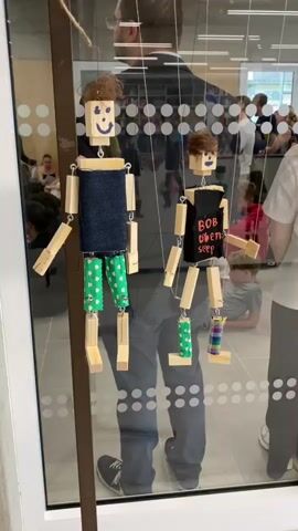 The video shows a display of several wooden puppets hanging behind a glass pane. The puppets are made of rectangular wooden blocks for the head, torso, and limbs, with smaller blocks or beads for the joints. They are suspended by strings attached to a horizontal bar above. The puppets are dressed in various colorful and patterned clothing made of fabric. Some of the puppets have painted faces with simple smiles. In the background, through the glass, a group of people can be seen, some standing and looking at the display, while others are seated on the floor. The environment appears to be indoors, possibly a school or community center, with a tiled floor and some shelving units visible. The lighting is bright, suggesting an indoor space with artificial or natural light. The camera is positioned at eye level, focusing on the puppets and the reflections in the glass. The video pans slightly to the right, revealing more of the display and the people in the background.