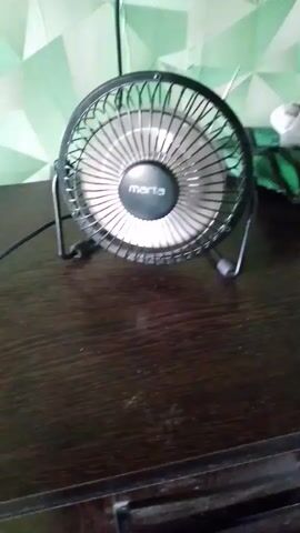 A black fan with a silver grille sits on a dark brown wooden surface. The fan has a circular head with blades visible behind the grille and the brand name "marta" printed on the front. It is supported by two black metal legs. Behind the fan, a green and white patterned wallpaper or fabric is visible. The fan appears to be stationary.