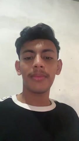 A young man with short black hair and a mustache is looking directly at the camera, talking. He is wearing a black t-shirt with white accents on the collar and shoulders. The background is a plain, off-white wall. The lighting is dim.