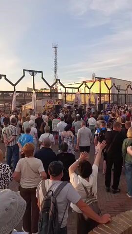A crowd of people stands gathered outdoors, facing a stage or performance area where a band appears to be playing. The sky is clear with a soft, warm light, suggesting it is late afternoon or early evening. The architecture in the background features a series of arched structures. People in the crowd are dressed casually, and some are raising their hands.