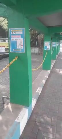 A stationary wide shot looks down a paved walkway lined with green pillars. A yellow chain is attached to each pillar, creating a barrier along the walkway. Several posters are affixed to the pillars, some featuring images and text in Indonesian. One poster depicts a person wearing a hard hat and holding a ball. To the left of the walkway, a larger billboard is visible, also displaying Indonesian text and images. Trees and foliage are present in the background. The scene appears to be outdoors, under daylight.