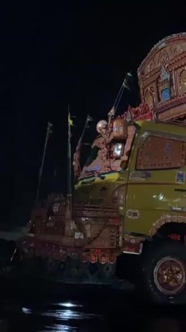 A nighttime exterior shot shows a heavily decorated truck with intricate designs and colorful lights on its exterior. The truck is yellow and brown. The side of the truck has a large cargo area adorned with detailed artwork and multiple lights. A person wearing a dark jacket and a head covering walks alongside the truck. The ground appears wet, suggesting it might have rained recently. The background is dark, indicating it is nighttime.