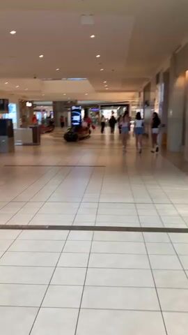 The video shows an interior view of a shopping mall. The floor is tiled in a grid pattern. Several people are walking in the distance. There are storefronts visible, including one with a sign that says "PICKLE BARREL". Some retail displays with green bins are also present. The lighting appears to be artificial.