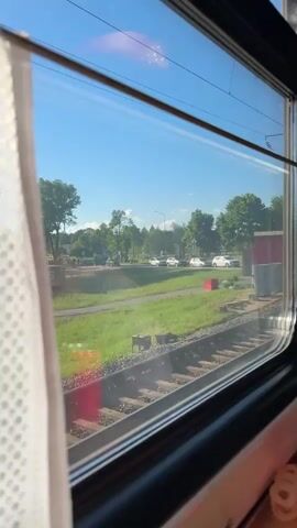 From inside a moving train, a view of the passing landscape is visible through a window. The foreground features train tracks, while beyond them lies a grassy area with trees and a street with cars. Buildings are also visible in the distance. A portion of a white, semi-transparent curtain hangs in the left side of the frame.