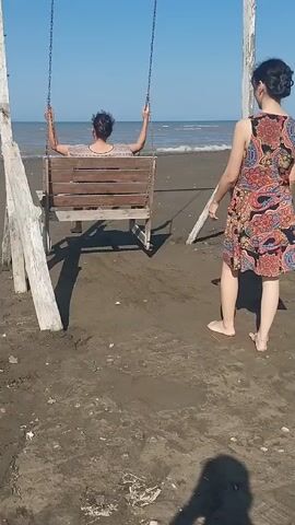 A video shows two women at a beach. One woman is seated on a wooden swing with a high back, facing the ocean and holding onto the swing chains. Her hair is dark and tied back. She is wearing a light-colored patterned top. The second woman, with dark hair also tied back, stands behind the swing, to the right of the seated woman. She is wearing a colorful, patterned dress and appears to be pushing or stabilizing the swing with her right hand. The beach sand is dark and appears wet in places, with some debris scattered on it. The ocean is visible in the background, with a clear blue sky above. Two weathered wooden posts support the swing. The overall lighting suggests it is daytime and sunny.