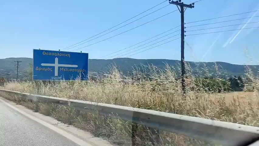 The video captures a car driving along a road on a sunny day. The camera is positioned from inside the car, looking out the front passenger window. Initially, a blue road sign with Greek text and an arrow indicating directions is visible on the left side of the road. The sign indicates directions to Thessaloniki, Drymos, and Melissochori, with "400M" written below. As the car continues, the sign moves out of view. The road appears to be a two-lane highway with a guardrail on the left. On the right side of the road, there's a field with tall, dry grass and some green bushes. Beyond the field, power lines with wooden poles stretch across the landscape. In the distance, hills covered with trees and some scattered buildings can be seen under a clear blue sky. A red triangular warning sign with a black exclamation mark is visible on the right side of the road for a moment. The car maintains a steady speed throughout the video.