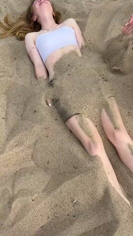 The image shows a young woman buried in sand up to her torso. She is lying on her back with her head tilted up, and her eyes are closed. She is wearing a white crop top and shorts. Her arms are also partially buried in the sand, with one hand visible on her left side. Her legs are exposed and extend out from the sand. The sand appears to be wet in some areas, and there are some debris visible on the surface. The lighting suggests it is daytime, likely sunny given the shadows. The scene appears to be a beach or a sandy area. A person's foot in red and white patterned shorts is visible in the lower left corner of the frame, suggesting someone is interacting with the buried woman. Another foot is visible in the lower right corner. The overall mood is playful and relaxed, indicating a fun activity on a sunny day. There is no visible text in the image.