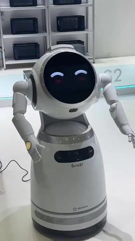 A white robot with a circular head displaying animated eyes is shown in a brightly lit exhibition space. The robot has two articulated arms with robotic hands. The robot's body is a white cylindrical base with grey accents. The robot's face displays a sequence of expressions: first a neutral face, then arms raised in a welcoming gesture, then a gesture with one arm extended and the other bent, and finally a gesture with one hand near its head. In the background, shelves are visible, stocked with black electronic devices. The floor is white.