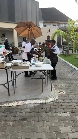The video shows an outdoor catering setup next to a swimming pool. People are swimming in the pool, and tables with umbrellas are set up around the pool area. There are people seated at the tables. A catering crew is setting up food on tables. One person appears to be preparing food on a vertical rotisserie. Another person is working at a table with various containers and plates. Servers are present, dressed in uniforms with aprons. The area is paved with gray bricks. Palm trees and green grass are also visible. The video is taken during daylight hours, with a clear sky overhead.