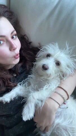 A woman with long, dark, curly hair, wearing a dark shirt, holds a small, white, scruffy-haired dog in her arms. She lies on a light-colored couch and looks at the camera, puckering her lips. The dog looks at the camera. The woman wears multiple bracelets on her right wrist.