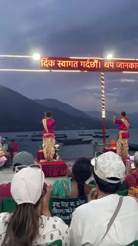 Two individuals are on small raised platforms in front of a lake, facing away from the camera and towards the water, performing what seems to be a synchronized dance or ritual. They are dressed in similar traditional attire with pink tops and cream-colored bottoms. Mountains are visible in the background, across the lake. A small crowd of people is seated in front of the performers, watching. Some are taking photos or videos with their phones. A sign in Nepali script is above the performers, illuminated by LED lights. Boats are scattered on the lake. The sky is overcast, suggesting it is either early morning or late evening.