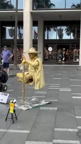The video shows a street performer dressed and painted entirely in gold, including their clothes, hat, and skin. They are sitting cross-legged atop a gold-colored pole, giving the illusion of levitation. The setup is placed on a silver-colored sheet on a pedestrian walkway in front of a building. To the left of the performer, there is a tripod stand with a sign that says "Tips." A young girl approaches the performer and shakes the performer's hand. A man stands nearby observing the scene. The surrounding environment includes palm trees, a modern building with glass doors, and other people passing by.