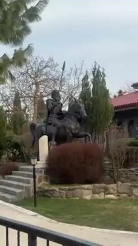 The video presents a collection of outdoor shots, likely taken in a historical or cultural site. The initial scene features a bronze statue of a medieval warrior on horseback, holding a spear, situated in a garden area with trees, shrubs, and stone steps. The camera then pans to reveal an arched structure constructed of red and beige bricks, adorned with inscriptions in a Cyrillic alphabet. On top of the arch, a statue depicting a saint or religious figure standing atop a globe is visible. Beneath the arch, another sculpture can be seen. The overall setting suggests a place of historical or religious significance, captured on a cloudy day. There are no visible people or audible sounds in the video.