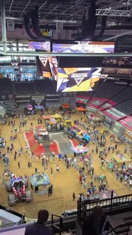 The video shows a high-angle view of a Monster Jam pit party event. Several large monster trucks are on display on a dirt arena floor. A bright blue and pink monster truck with the branding "Bailey Circuit" is prominently featured in the foreground, with a matching tent set up nearby. Other monster trucks, including a yellow one and an orange and red one, are also visible. Spectators, including families and children, are walking around the arena, interacting with the trucks and the event. Tents and booths are set up throughout the area, suggesting merchandise and other activities. A large screen displaying the Monster Jam logo is visible in the background, along with stadium seating. The overall atmosphere appears to be lively and family-friendly.