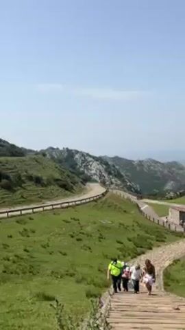 The video shows a panoramic view of a mountainous landscape on a sunny day. In the first shot, people are walking on a stone path near a road. The road has a guardrail, and the path leads to a green hillside with trees. As the camera moves, it captures more of the landscape, revealing rolling hills, grassy areas, and stone structures that appear to be ruins. The sky is blue with a few scattered clouds. The overall atmosphere suggests a scenic and historical location.