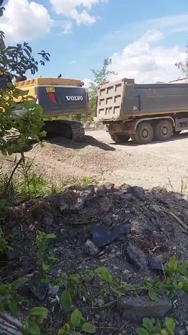 An excavator is loading a dump truck at a gravel pit. The excavator is yellow and black and has the Volvo logo on it. The dump truck is dark gray. The scene is outdoors on a sunny day with some clouds in the sky. There are trees and bushes in the foreground. The ground is covered in gravel and dirt. The excavator is in the process of digging into a pile of gravel.