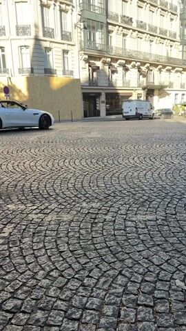 A white convertible sports car drives on a cobblestone street in front of a building. A white van is parked to the right of the sports car. There are other cars in the background. The sun is shining.