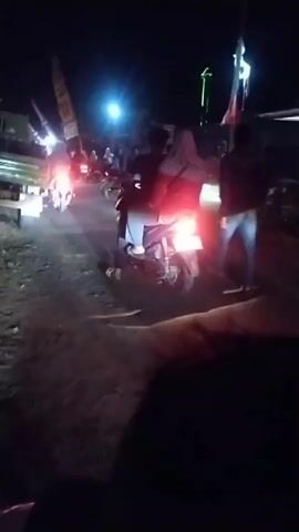 A nighttime scene on a dirt road shows several people and motorcycles. In the foreground, the camera is held low, possibly by someone sitting on a motorcycle. A motorcycle with a rider wearing a hijab and a red jacket is in the center of the frame, its taillight illuminated. Another person is visible behind the rider. To the left, a truck with its brake lights on is partially visible, with other motorcycles and people around it. The background is dark, with some lights and structures suggesting a rural or semi-urban environment. The ground is uneven and dusty.