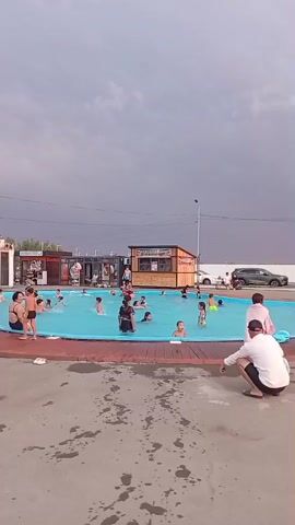 A long shot captures a bright blue swimming pool filled with people of various ages enjoying themselves under a cloudy sky. Several children are actively swimming and playing in the water. On the poolside, a man wearing a white shirt and black shorts crouches down, facing the pool. To his right, a person in a light pink robe stands looking towards the water. In the background, a small building with a wooden facade and a sign is visible, along with another structure with signage. Cars are parked beyond the pool area. The ground surrounding the pool is made of concrete, with visible wet patches.