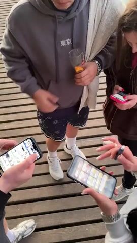 The video shows a group of four people standing on a wooden pier, each holding a smartphone. They are looking at their phones. One person is holding a glass with a drink, and another has a towel draped over their shoulder. The pier is made of planks and is located near a body of water. The people are casually dressed in hoodies, shorts, and sneakers.