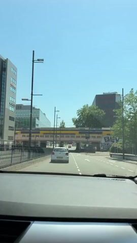 The video shows a view from inside a car driving down a road in Eindhoven, Netherlands. The car is following a silver Volkswagen Polo. Above the road, a yellow and blue train passes by on an elevated railway track. The train appears to be a Dutch Railways (NS) Intercity train.

The scene is set during the daytime, with a clear blue sky and bright sunlight, indicating fair weather. Modern buildings, one with a glass facade, are visible on the left side of the road. On the right side, there's a building with red accents and what appears to be graffiti on a wall beneath the railway bridge. Green trees are also present in the background.

Notable details include a speed limit sign indicating "40m" (likely 40 kilometers per hour, as it's a common speed limit in Dutch urban areas, and the 'm' might be a typo or part of a larger sign). The license plate of the car in front is visible: "K-402-NG". The graffiti on the wall beneath the bridge has visible text, including what looks like "BEV" and other illegible markings. There are streetlights and power lines visible throughout the scene. The overall impression is of a typical urban driving experience in a European city on a sunny day.
