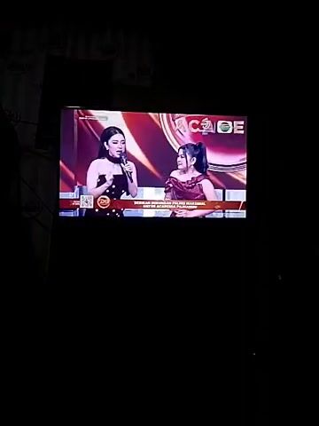 A television screen displays two women on a stage during a broadcast. The woman on the left wears a black, polka-dotted dress and holds a microphone. The woman on the right wears a burgundy dress with off-the-shoulder sleeves. Both women are looking at each other, with the woman on the left appearing to be speaking. The background on the screen is a reddish-orange hue with the word "ACADE" prominently displayed in gold letters. Text in smaller font is visible at the bottom of the screen, indicating a program title and potentially other broadcast information. The overall environment is dark, suggesting the image was taken in a dimly lit room.