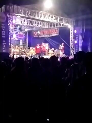 A nighttime outdoor concert is in progress. A stage is set up with a band performing. The band members are on stage, some wearing red shirts. A banner with "MURENSA" is displayed above the stage. A crowd of people is gathered in front of the stage, silhouetted against the bright stage lights. The crowd appears to be enjoying the performance. The environment is dark, illuminated primarily by the stage lights.