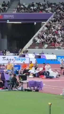 The video shows a pole vaulter making a jump at a sporting event. The vaulter is wearing a blue and yellow uniform. The pole vault bar is raised high and supported by two purple vertical posts.  The vaulter runs down the track with the pole and plants it in the vaulting box, propelling him upward. He successfully clears the bar and lands on a padded mat. The audience, visible in the background, cheers. Several people and cameras are seen along the side of the track. A digital clock is seen at the right side of the video at the edge of the landing pad, displaying the time "22:18:37".  The event appears to be held in a large stadium with numerous spectators present. The lighting is bright, indicating an outdoor event.