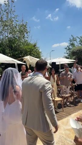 A wedding ceremony is underway outdoors. A bride in a white wedding dress and veil walks with a groom in a light-colored suit. They are walking down an aisle made of wooden planks, with guests seated on either side. The guests are dressed in casual attire, and many are holding up their phones to take pictures or videos. Some guests are clapping and cheering as the couple walks by. The environment is sunny with a blue sky and some clouds. There are trees and white canopies in the background, suggesting an outdoor venue such as a garden or patio. The overall atmosphere appears celebratory and joyful.