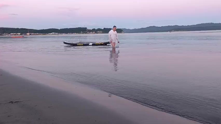A man in a white shirt and shorts walks in shallow water near a sandy beach. He is pulling a paddleboard. The sky is a soft pink and purple, suggesting dawn or dusk. Hills and a distant coastline are visible across the water. The paddleboard has lettering in yellow.