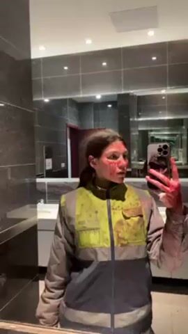 A young woman stands in front of a bathroom mirror, taking a selfie with a phone held in her right hand. Her face and parts of her neck are covered in what appears to be fake blood, as are her right hand and the yellow and gray safety jacket she is wearing. The bathroom has dark gray tiled walls and multiple mirrors, reflecting the scene. The lighting is bright, and the overall impression is of a staged or costumed appearance.