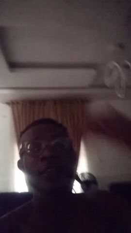 A person wearing glasses is visible in a dimly lit room. The person raises a hand up in the air in front of a window with closed curtains. The room has a white ceiling.