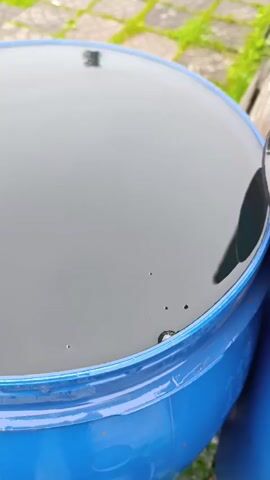 A close-up video shows the surface of black liquid in a blue plastic barrel. Bubbles rise to the surface in a few spots near the rim of the barrel. The barrel is outside on a paved surface with some green foliage visible in the background.