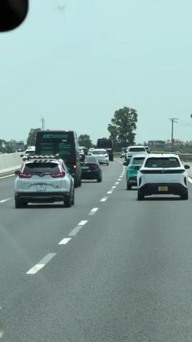 The video shows the rear view of several vehicles moving down a highway during daytime. The sky is overcast. Visible vehicles include a silver Honda CRV, a dark van bearing the logo "DC LANDSCAPE", and a white VinFast VF8. The vehicles are moving at a moderate speed in a line. The road is marked with white dashed lines separating the lanes. Green foliage and utility poles are visible on the side of the highway. The video is taken from inside another moving vehicle.