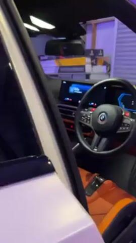 The video shows the interior of a parked BMW. The car has black trim, orange seats, and red ambient lighting. The steering wheel is black with the BMW logo in the center. There are also screens displaying information on the dashboard. The car is parked in a garage.