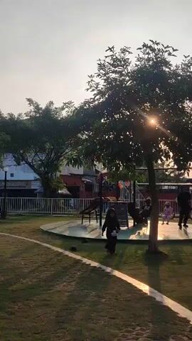 The video shows a park scene during sunset. The sun is low in the sky, casting a warm glow and creating lens flare through a tree in the foreground. A woman in a hijab is walking on the grass. In the background, there is a playground with children and adults. Some people are sitting on a bench under a tree. The park has a grassy area, a paved path, and playground equipment.