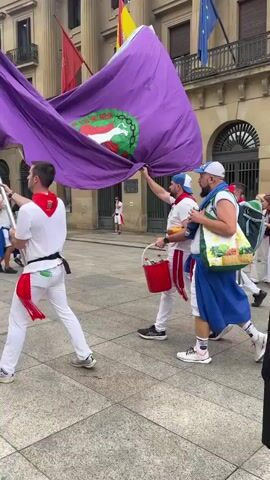 The video shows a group of people walking in what appears to be a parade or procession. They are on a paved square in front of a large building with classical architecture. Many of the participants are wearing white clothing with red sashes or bandanas, and some are wearing blue waist sashes. Two large purple flags with green emblems are being carried aloft by several individuals. One person is carrying a red bucket. Another person is carrying a tote bag with an image of produce on it. In the background, there are flags of Spain and the European Union displayed on the building. Some participants are playing musical instruments, including a trombone. The atmosphere appears festive.
