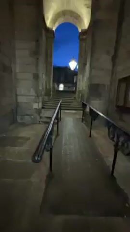 The video shows a passage under an arched stone structure. The passage has railings on either side leading up to a set of steps. The sky is visible through the archway, and it appears to be twilight. The arches of the structure are illuminated with warm lighting, and there are additional lights hanging from the ceiling within the structure. As the camera pans around, it shows a view of the street outside the archway, with buildings and lights visible in the distance. There are also some bicycles parked nearby. The overall impression is that it is a historic building with impressive architecture.